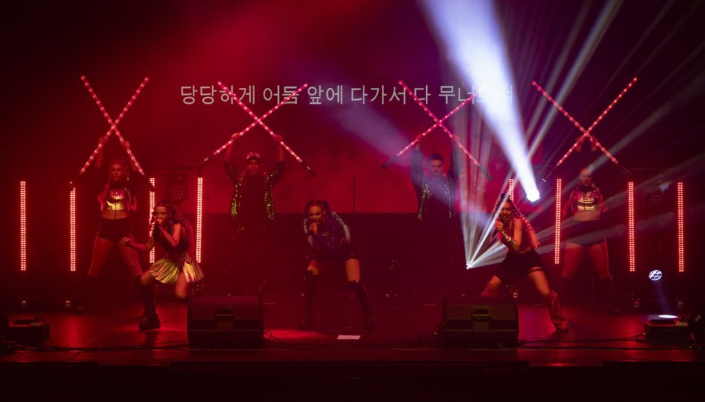 K-Pop Fantasy cast performing with light sticks under red stage lighting