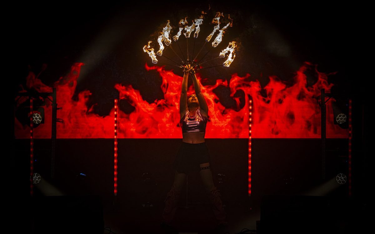 A K-Pop Fantasy performer raises a flaming fan in front of a wall of fire visuals