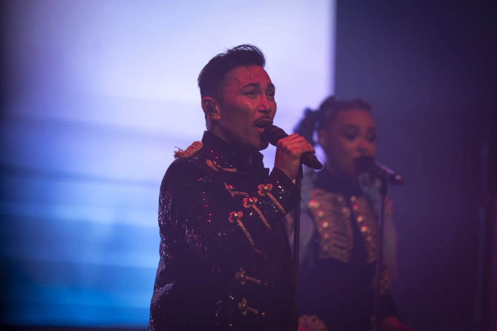 A K-Pop Fantasy singer in a sequinned jacket performs in front of the screen